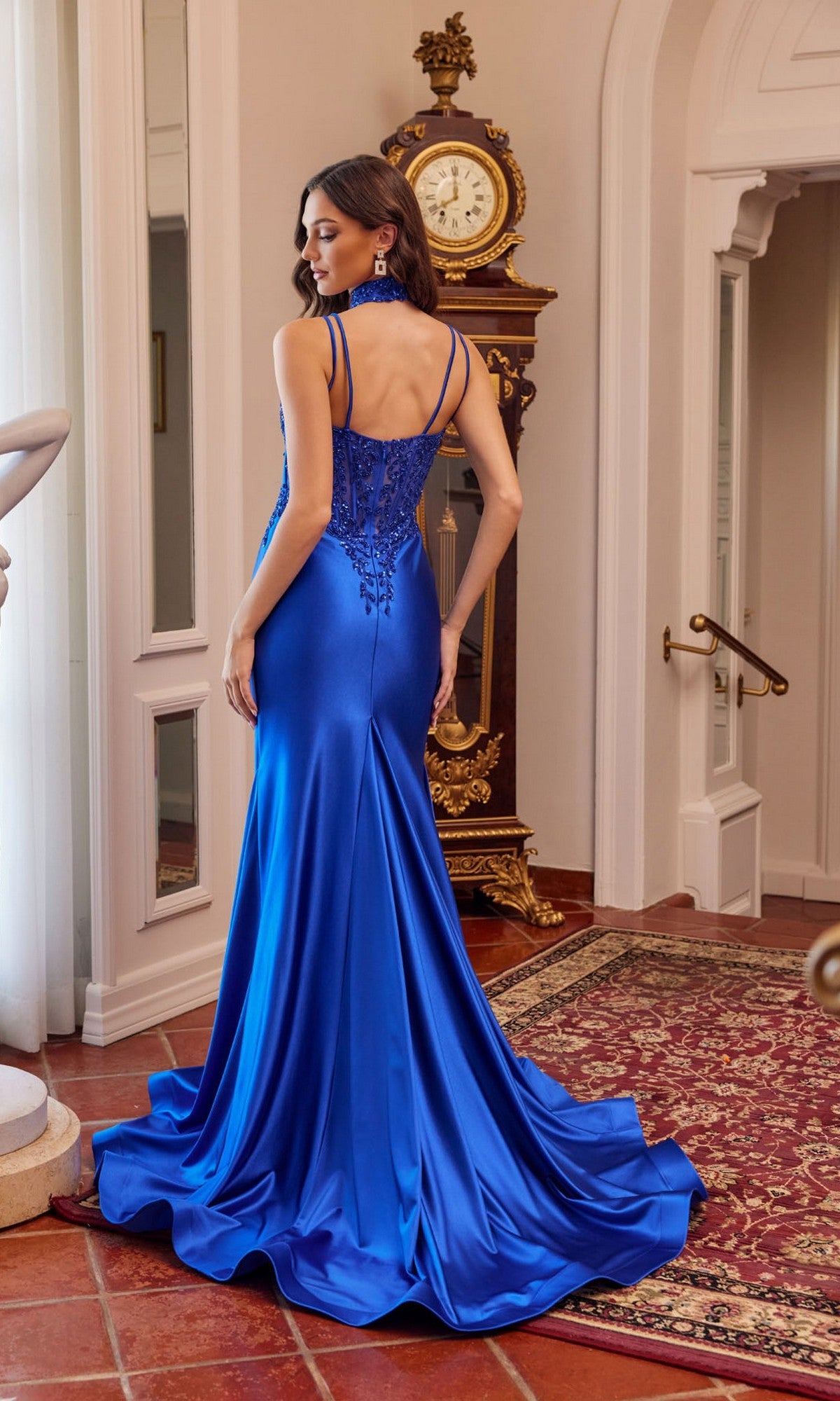 A woman wears the Juliet JT2628L long royal blue satin prom dress with a sweetheart neckline and delicate embroidery, standing in an elegant room with a grandfather clock and red patterned rug.